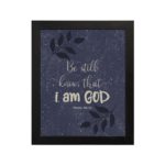 Purple-blue background with black leaf design, white text Be Still Know That I Am God Psalm 46:10 Bible Verse Wall Art black frame hung on the wall.