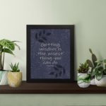 Christian wall art with purple-blue background, black leaf design, white font displaying Proverbs 4:7 Bible Verse, and black frame on the table