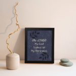 Purple-blue background with black leaf design, white text 'The Lord My God Lights Up My Darkness Psalm 18:28 Bible Verse Wall Art black frame displayed on the wall.