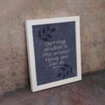 Christian wall art with purple-blue background, black leaf design, white font displaying Proverbs 4:7 Bible Verse, and white frame on the wall