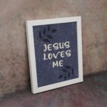 Purple-blue background with black leaf design, white text Jesus Loves Me Bible Verse Wall Art white frame on the wall.