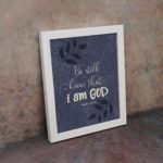 Purple-blue background with black leaf design, white text Be Still Know That I Am God Psalm 46:10 Bible Verse Wall Art white frame on the wall.