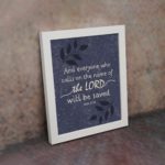 Christian wall art with purple-blue background, black leaf design, and white text And Everyone Who Calls On The Name Of The Lord Will Be Saved Acts 2:21 Bible Verse Wall Art white frame on the wall