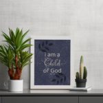 Christian wall art with purple-blue background, black leaf design, and white text I Am A Child Of God John 1:12 Bible Verse Wall Art white frame on the table