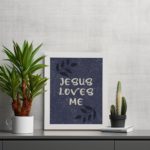 Purple-blue background with black leaf design, white text Jesus Loves Me Bible Verse Wall Art white frame on the table.