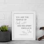 Christian wall art featuring the verse You Are The Temple Of God, His Spirit Dwells In You from 1 Corinthians 3:16 on a gray background with gray leaf design and black and gray font white frame displayed on the table.