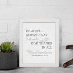 Christian wall art featuring 1 Thessalonians 5:16-18 Bible verse on a white background with gray leaf design and black font and black frame displayed on the wall