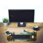 Desk pad featuring a serene landscape of mountains, pine trees, and a calm lake with a soft blue and green color palette. Desk Pad with Keyboard and Mouse on the table