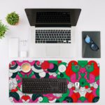 Colorful abstract desk pad with playful patterns, featuring pink, green, red, black, and white shapes resembling flowers and circles. Desk Mat with an Apple Laptop