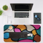 Infidu Artistic Desk Mat with organic, curving multicolored shapes in pink, light blue, yellow, gray, and white. Desk Mat with an Apple Laptop