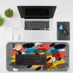 Infidu Artistic Patterns Desk Pad with bold, brushstroke-like shapes in yellow, black, red, and white on a gray background. Desk Mat with an Apple Laptop