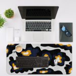 Infidu Artistic Patterns Desk Pad with a black background and large, irregular white and orange abstract shapes. Desk Mat with an Apple Laptop