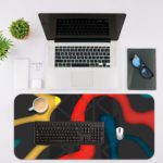 Infidu Artistic Desk Mat with bold curvy shapes in yellow, red, and blue on a dark black background. Desk Mat with an Apple Laptop