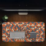 Desk pad with an abstract geometric pattern in blue, orange, and white, featuring triangles and various shapes. Desk Pad in computer table