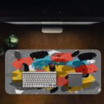 Infidu Artistic Patterns Desk Pad with bold, brushstroke-like shapes in yellow, black, red, and white on a gray background. Desk Pad on computer table