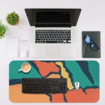 Infidu Artistic Desk Pad with angular shapes in green, orange, and blue on a dark background. Desk Mat with an Apple Laptop