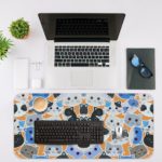 Desk pad with colorful blue, grey, and black cat faces on a sandal-colored background. Desk Mat with an Apple Computer