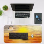 Desk mat featuring a sunset scene with a silhouette of a sailboat on an orange-gold sea, under a warm yellow and golden sky. Desk Mat with an Apple Laptop