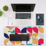 Infidu Artistic Desk Mat with a colorful geometric pattern of overlapping circles and quarter-circles in navy, pink, orange, teal, and yellow. Desk Mat with an Apple Laptop