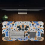 Desk pad with colorful blue, grey, and black cat faces on a sandal-colored background. Desk Pad with Design Kept on a computer table