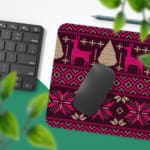 Infidu Desk Pad with a deep purple background and bright pink and white knitted pattern featuring reindeer and trees. Design Kept on a computer table