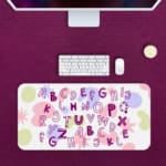 Infidu Alphabet Desk Pad with a white background featuring colorful letters and star shapes in various styles and colors. Desk Pad with Keyboard and Mouse in pink background