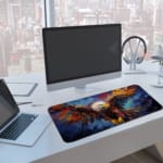 Colorful eagle with spread wings, white head, brown body, and vivid red and orange feathers against a blue abstract background. A computer and laptop are next to the desk pad