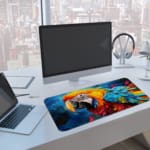 A bright parrot with vibrant feathers against a dynamic blue background. A computer and laptop are next to the desk pad