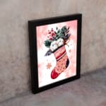 Christmas wall art featuring a pink stocking with red accents, filled with green leaves, berries, and white snowball-like objects, on a light pink background with star patterns, framed in black display on the wall in a black frame