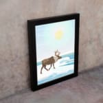 Christmas wall art featuring a reindeer walking on snow near a body of water with a bright sun and blue sky in the background. on the wall in a black frame