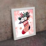 Christmas wall art featuring a pink stocking with red accents, filled with green leaves, berries, and white snowball-like objects, on a light pink background with star patterns, framed in black display on the wall in a white frame