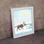 Christmas wall art featuring a reindeer walking on snow near a body of water with a bright sun and blue sky in the background. on the wall in a white frame