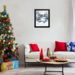 Infidu A Joyous Holiday Wall Art - Tranquil Winter Forest Scene with Deer shows a snowy forest with evergreen trees, a blue house, and a deer. hung on the wall in a black frame