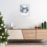 Infidu A Joyous Holiday Wall Art - Tranquil Winter Forest Scene with Deer shows a snowy forest with evergreen trees, a blue house, and a deer. hung on the wall in black frame in a white frame