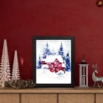 Christmas wall art featuring a pink house and blue pine trees in a watercolor style with a light background. displayed on the table in a black frame