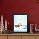 Christmas wall art featuring a reindeer walking on snow near a body of water with a bright sun and blue sky in the background. displayed on the table in a black frame