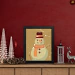 Snowman illustration with a red hat and orange scarf, framed in black, with snowflakes on a light beige background. displayed on the table in a black frame