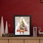 Christmas wall art featuring a white tree with red ornaments, gifts underneath, a reindeer, and a snowy background in a watercolor style. displayed on the table in black frame