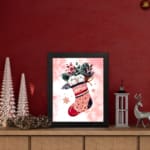 Christmas wall art featuring a pink stocking with red accents, filled with green leaves, berries, and white snowball-like objects, on a light pink background with star patterns, framed in black on the table