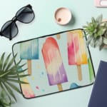 Laptop sleeve with pastel-colored popsicle design in blue, pink, purple, and peach on a light background with zip closure. the laptop sleeve on the table