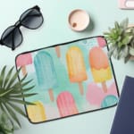 Laptop sleeve with light blue background and pastel popsicle pattern in pink, yellow, blue, and purple, featuring a zip closure. the laptop sleeve on the table