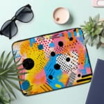 Laptop sleeve with vibrant abstract design in yellow, blue, pink, and black with circles, lines, and dots. the laptop sleeve on the table