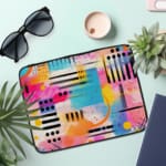 Infidu Bold Abstract Laptop Sleeve featuring bright geometric patterns in pink, blue, yellow, black, and white with a secure zip closure. the laptop sleeve on the table