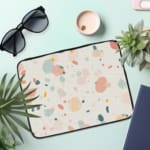 Pastel terrazzo-style laptop sleeve with pink, yellow, blue, and gray shapes on a light background, the laptop sleeve on the table