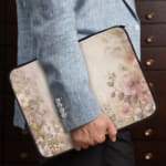 Soft beige laptop sleeve with delicate floral design in pink, green, and purple, featuring a zip closure for secure storage. the person holding the sleeve