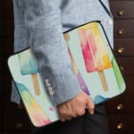 Laptop sleeve with pastel-colored popsicle design in blue, pink, purple, and peach on a light background with zip closure. the person holding the sleeve