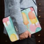 Laptop sleeve with light blue background and pastel popsicle pattern in pink, yellow, blue, and purple, featuring a zip closure. the person holding the sleeve