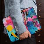 Laptop sleeve with a vibrant abstract design featuring blue, pink, purple, yellow, and black hues, secured with a zip closure. the person holding the sleeve