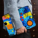 Laptop sleeve with a bright blue background featuring colorful abstract geometric shapes in yellow, pink, orange, black, and red, with zip closure. the person holding the sleeve