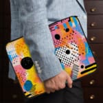 Laptop sleeve with vibrant abstract design in yellow, blue, pink, and black with circles, lines, and dots. the person holding the sleeve